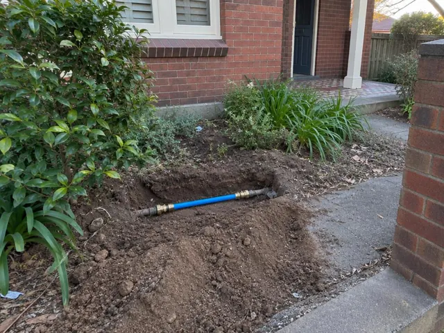 Open trench with a repaired blue water pipe beside a brick house and garden in Balgowlah